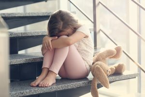 A young girl sitting on stairs with her arms wrapped around her knees and her head resting on her arms, next to a stuffed teddy bear, looking sad.
