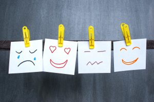 A row of four white pieces of paper, each with a different hand-drawn emoticon in various colors, clipped to a dark string with yellow clothespins. The first emoticon shows a sad face with tears, the second a happy face with heart-shaped eyes, the third a neutral face, and the fourth a big smile with closed eyes.