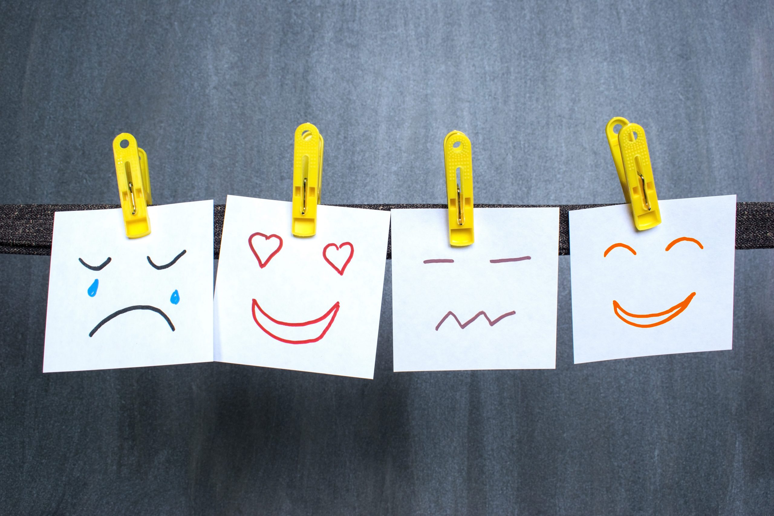 A row of four white pieces of paper, each with a different hand-drawn emoticon in various colors, clipped to a dark string with yellow clothespins. The first emoticon shows a sad face with tears, the second a happy face with heart-shaped eyes, the third a neutral face, and the fourth a big smile with closed eyes.