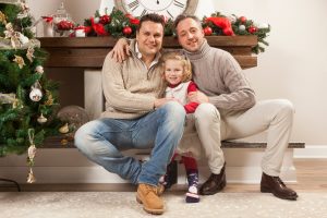 A family of three—two men and a young girl—sitting together on the floor in front of a fireplace decorated with red ornaments and greenery, smiling at the camera.