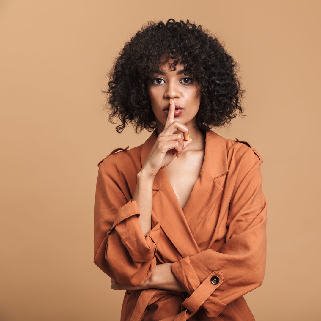 A woman with curly dark hair and medium skin tone is standing against a beige background. She is wearing a rust-colored blazer with rolled-up sleeves and has her left arm crossed over her chest. She is holding her right index finger up to her lips in a "shh" gesture, suggesting silence or secrecy.