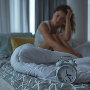 A lady sitting on the bed and a clock on the bed in front of her white blanket.