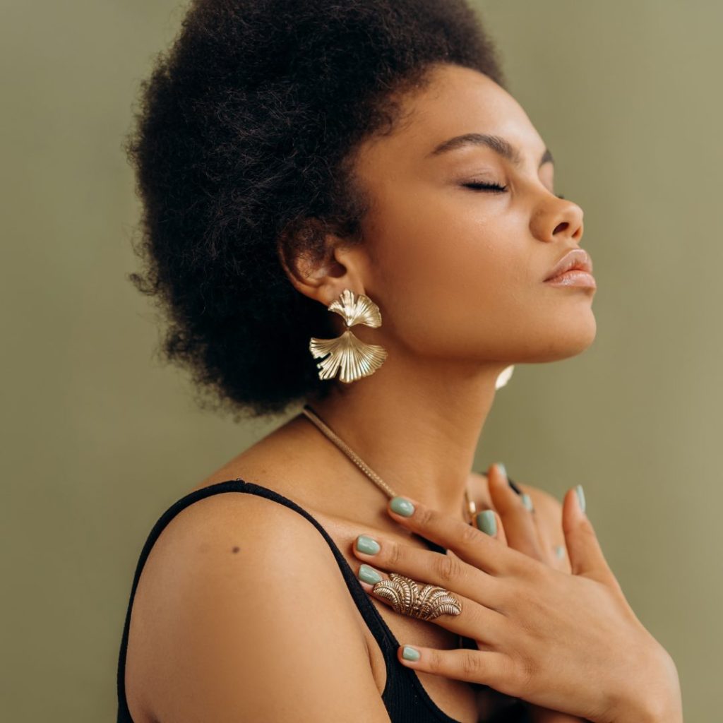 A woman with short, curly hair and medium skin tone has her eyes closed and appears to be in a peaceful moment. She is wearing large, gold, fan-shaped earrings and a delicate necklace. Her right hand, adorned with a large, ornate ring, is resting gently on her chest. She is dressed in a black spaghetti strap top, and the background is a soft, muted green.