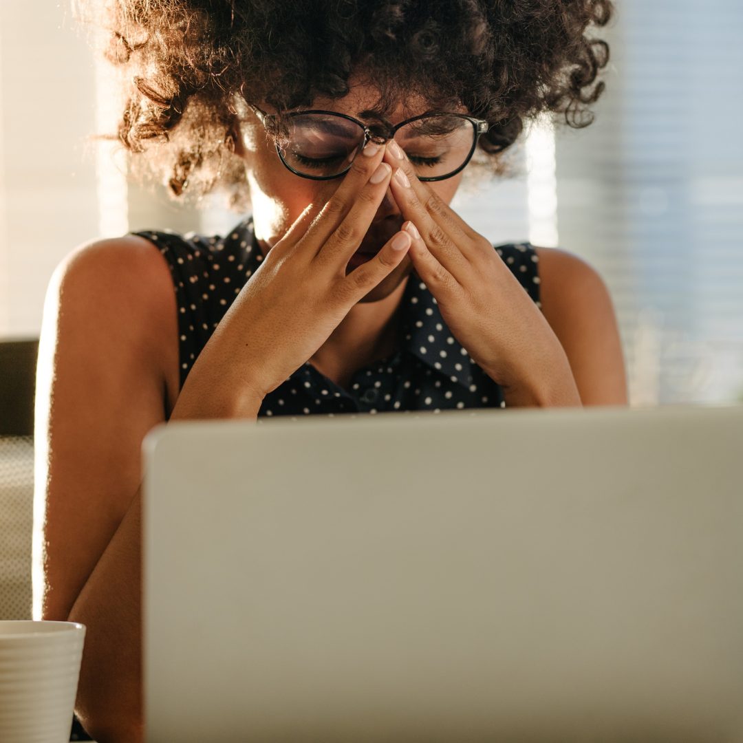 A woman with curly hair and glasses sitting at a desk with a laptop, holding her forehead with both hands and appearing stressed or overwhelmed. Sunlight streams through window blinds behind her.