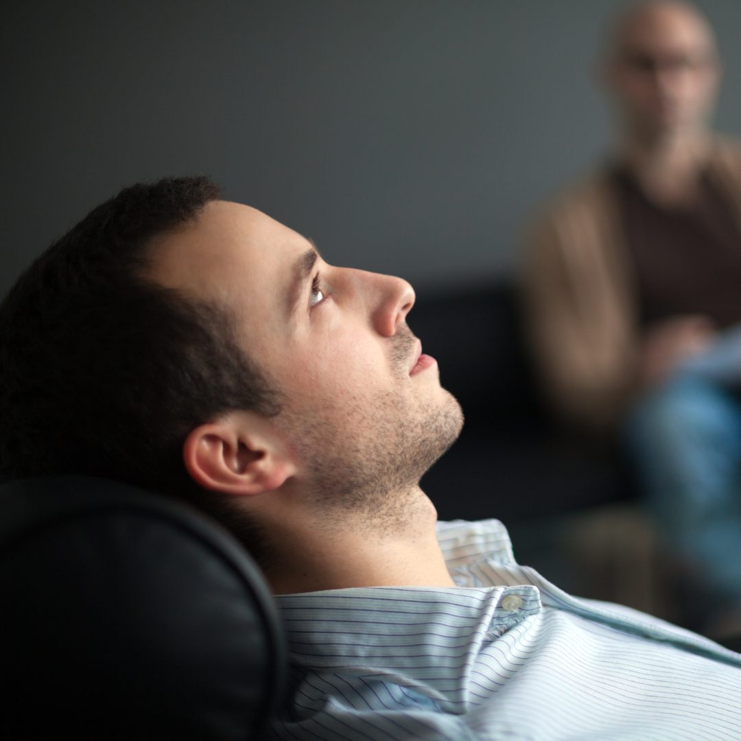 A man with dark hair and light skin is lying back in a chair, looking upwards with a thoughtful expression. He is wearing a light-colored, collared, striped shirt. In the background, there is a blurred figure of another person with glasses, dressed in brown, sitting on a dark-colored sofa. The setting appears to be indoors, possibly in an office or waiting room.