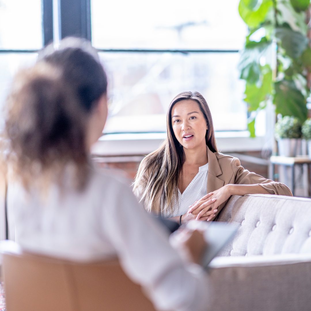 A lady with long hair sitting down and talking to a therapist.