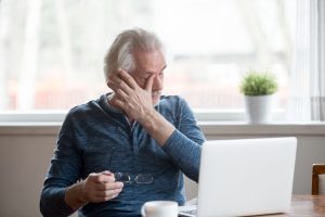 Older man sitting at a desk rubbing his eyes in fatigue while holding his glasses, with an open laptop in front of him.