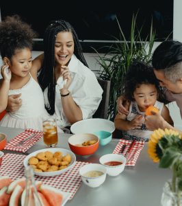 A family is gathered around a table enjoying a meal together. There are two young girls, one with curly hair and the other with braided hair, sitting next to a woman with long braided hair, all smiling and sharing a joyful moment. The table is set with various dishes including fried chicken nuggets, bowls of sauces, and slices of watermelon, with a glass of iced tea and a vase of sunflowers in the foreground. The background features green plants and a dark wall.
