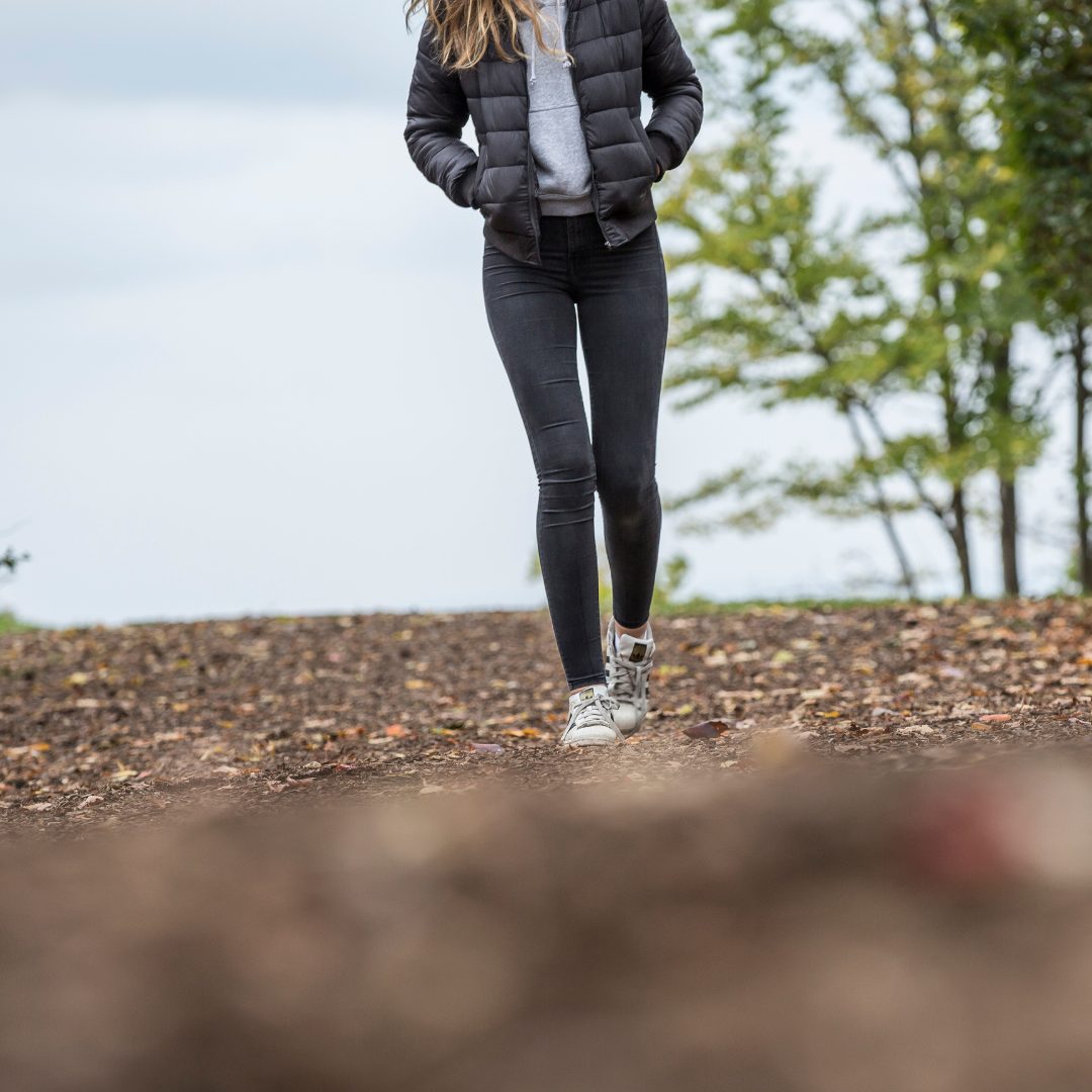 A lady wearing a black jacket and jeans with hands in her pocket walking.
