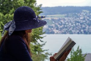 Woman wearing a wide-brimmed blue hat reading a book outdoors, overlooking a scenic lakeside landscape with distant hills and houses.