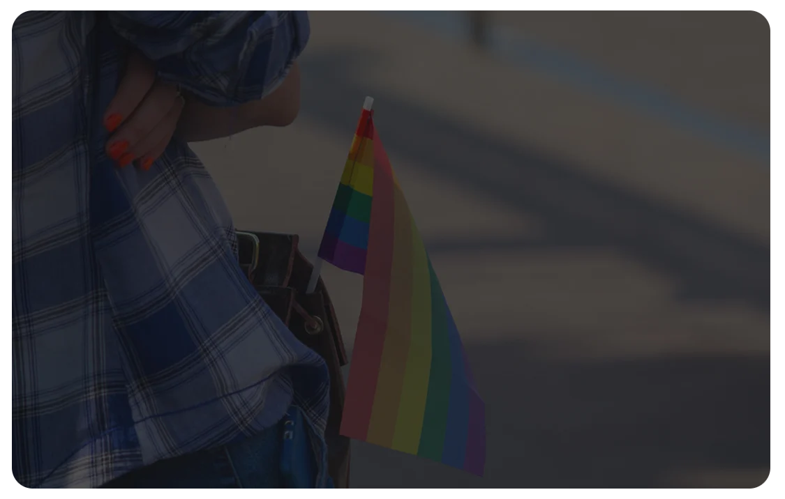 A person wearing a plaid shirt stands outdoors with a small rainbow pride flag attached to their bag, the colorful flag visible against a softly blurred background.