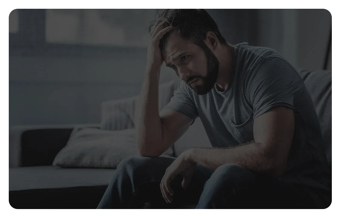 A bearded adult man sits on a couch in a dimly lit living room, leaning forward with his elbow on his knee and his hand pressed to his forehead, appearing stressed, worried, or deep in thought.