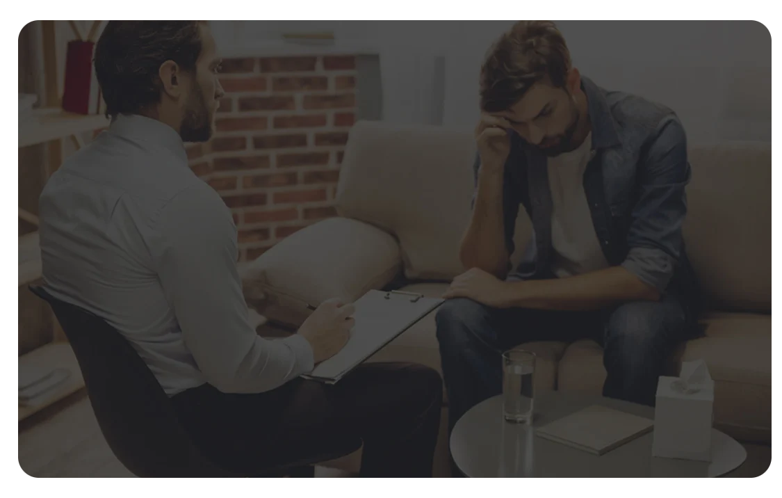 A man sits on a couch with his head resting on his hand, appearing distressed, while another man—likely a therapist—sits nearby holding a clipboard during a counseling session in a quiet office setting.