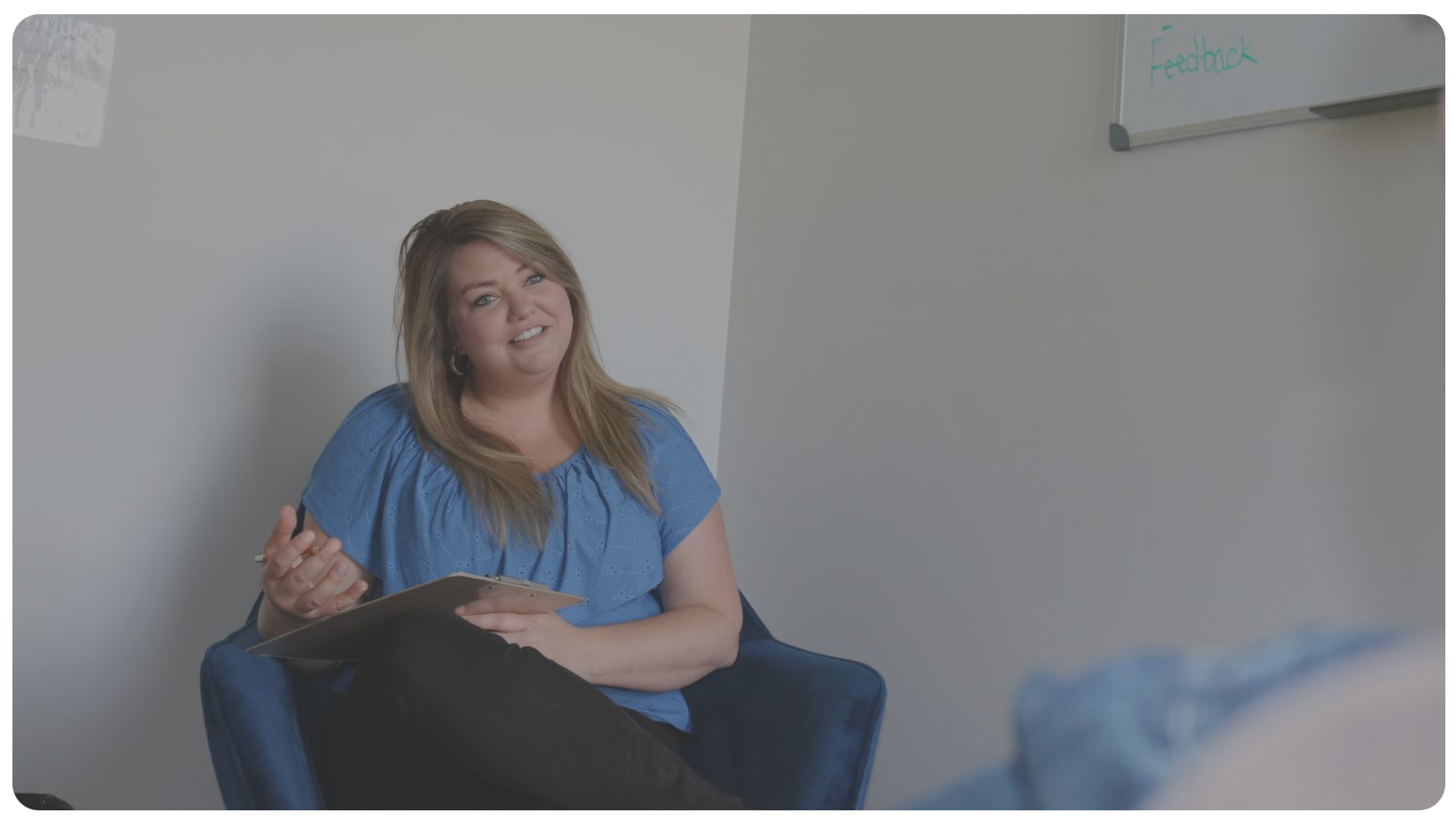 Breanne smiling, sitting down, wearing a green blouse, holding a pen and a clipboard.