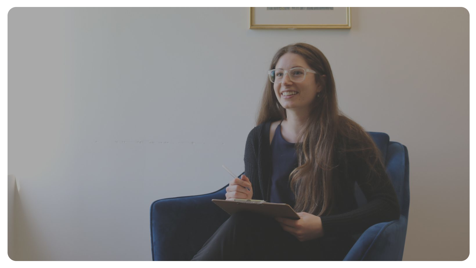 Jenny smiling, sitting down, holding a pen and a clipboard.