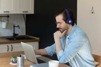 A man with glasses and dark hair, wearing headphones and a light denim shirt, sitting at a wooden table in a kitchen, looking at a laptop with his hand resting on his chin.