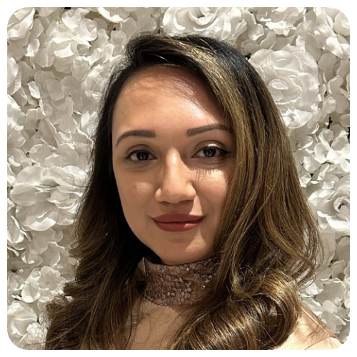 Jumana Daibl smiling, with long brown hair, wearing a light pink lace top, standing in front of a white floral background.