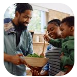 A man with dreadlocks and a beard smiling while holding a bowl of salad, engaging happily with a woman and a young child in a bright, cozy kitchen. The woman is smiling and holding a wooden spoon, while the child is reaching towards the bowl with curiosity. A man with dreadlocks and a beard smiling while holding a bowl of salad, engaging happily with a woman and a young child in a bright, cozy kitchen. The woman is smiling and holding a wooden spoon, while the child is reaching towards the bowl with curiosity.