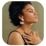 A woman with short, curly hair and medium skin tone stands against a muted green background with her eyes closed in a calm, reflective moment. She wears gold fan-shaped earrings, a delicate necklace, and a black spaghetti-strap top, resting a ring-adorned hand gently on her chest. A woman with short, curly hair and medium skin tone stands against a muted green background with her eyes closed in a calm, reflective moment. She wears gold fan-shaped earrings, a delicate necklace, and a black spaghetti-strap top, resting a ring-adorned hand gently on her chest.