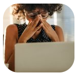 A woman with curly hair and glasses sitting at a desk with a laptop, holding her forehead with both hands and appearing stressed or overwhelmed. Sunlight streams through window blinds behind her. A woman with curly hair and glasses sitting at a desk with a laptop, holding her forehead with both hands and appearing stressed or overwhelmed. Sunlight streams through window blinds behind her.